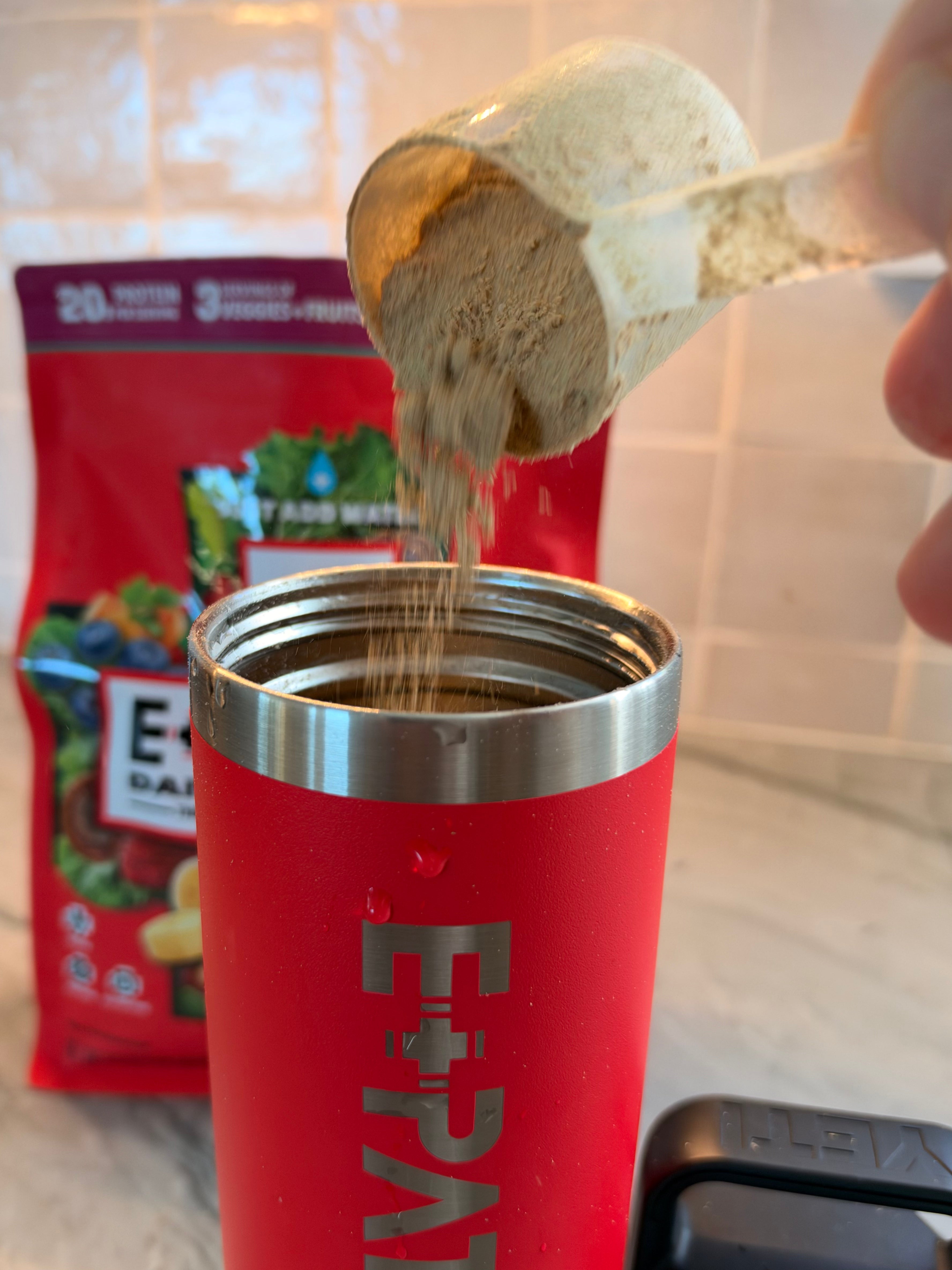 Scoop of E+PATROL powder being poured into a red Yeti waterbottle beside the pouch