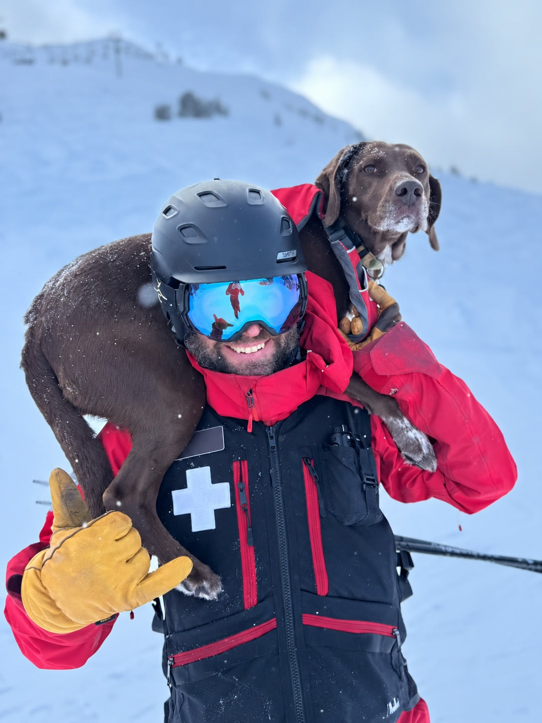 Ethan in his ski patroller gear (red rescue jacket), carrying a brown avalanche dog on his shoulders