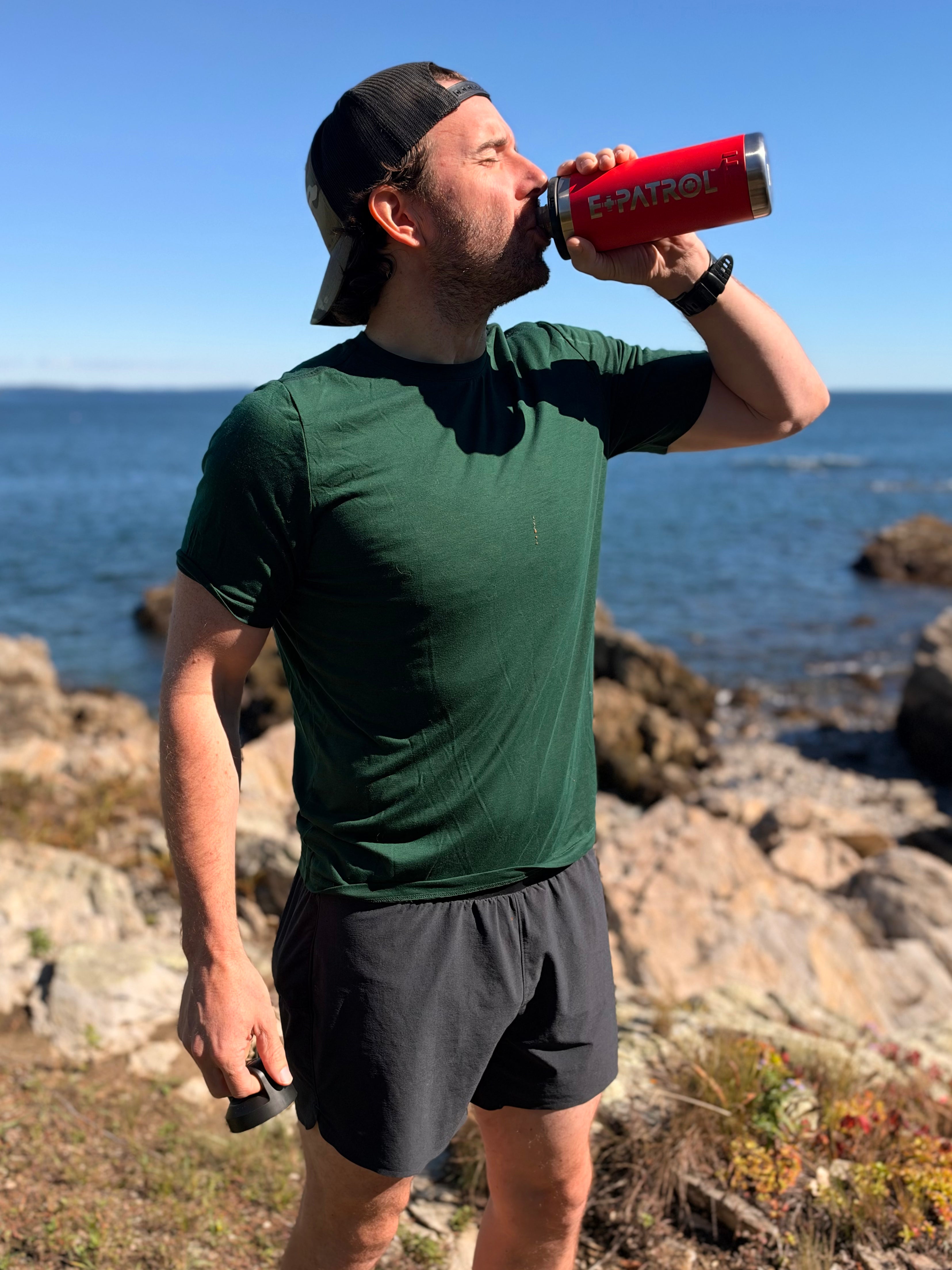 Man drinking from a red E+PATROL Yeti water bottle on a rocky shoreline